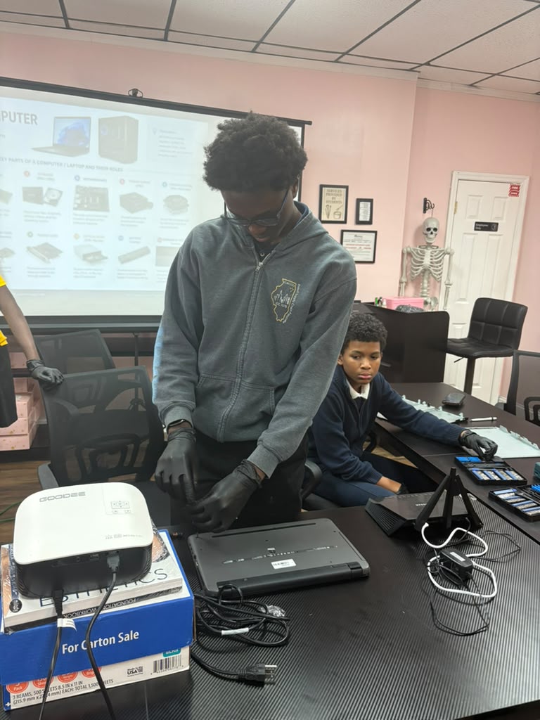 Two students working at a table with laptops and hardware components under projected architecture diagrams